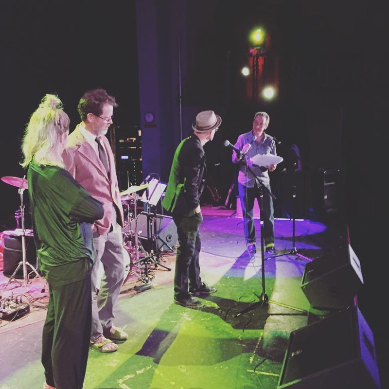 On stage at a theatre, a reader at the mic, 2016